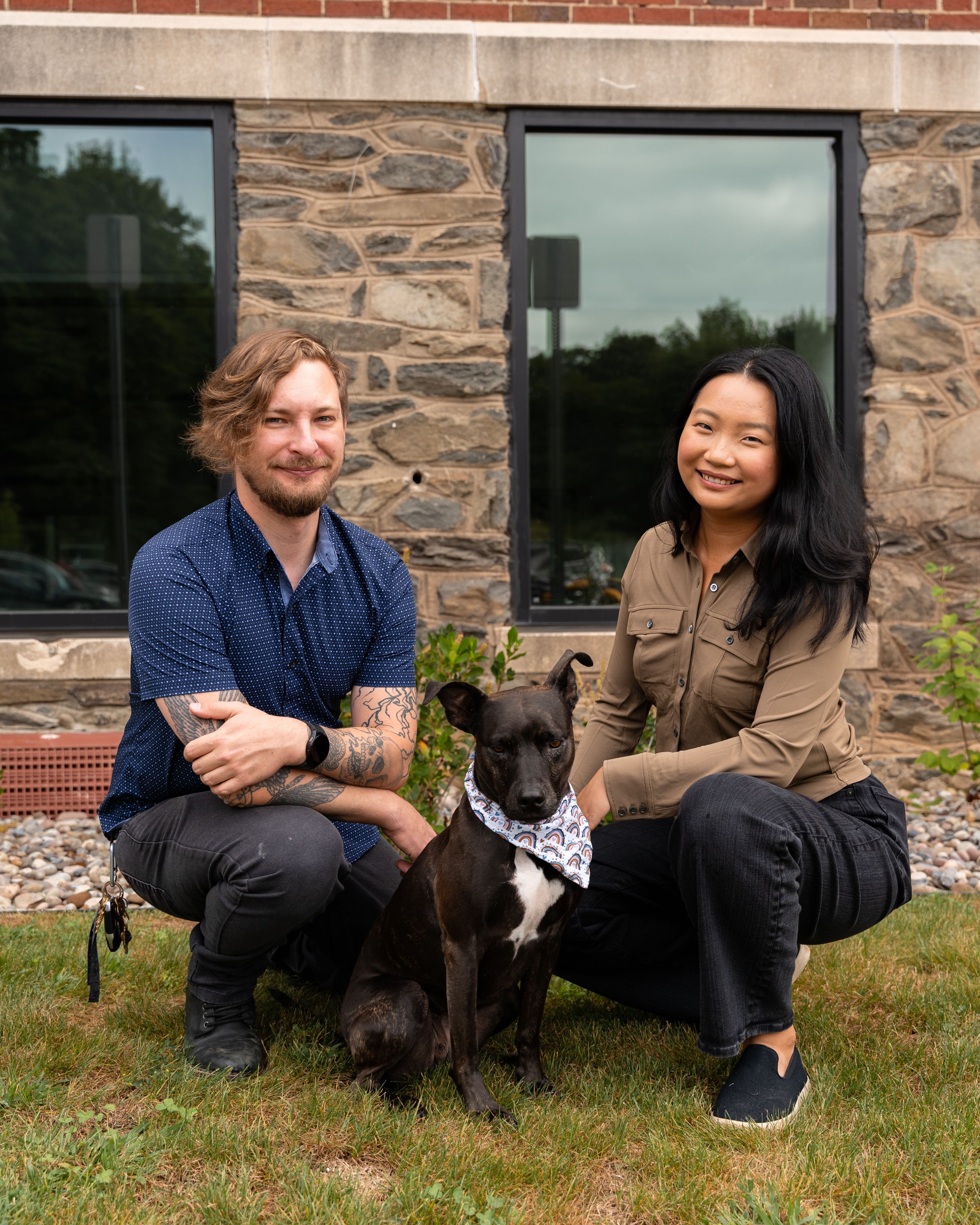 Two people sitting on grass with a dog in front of a stone building.