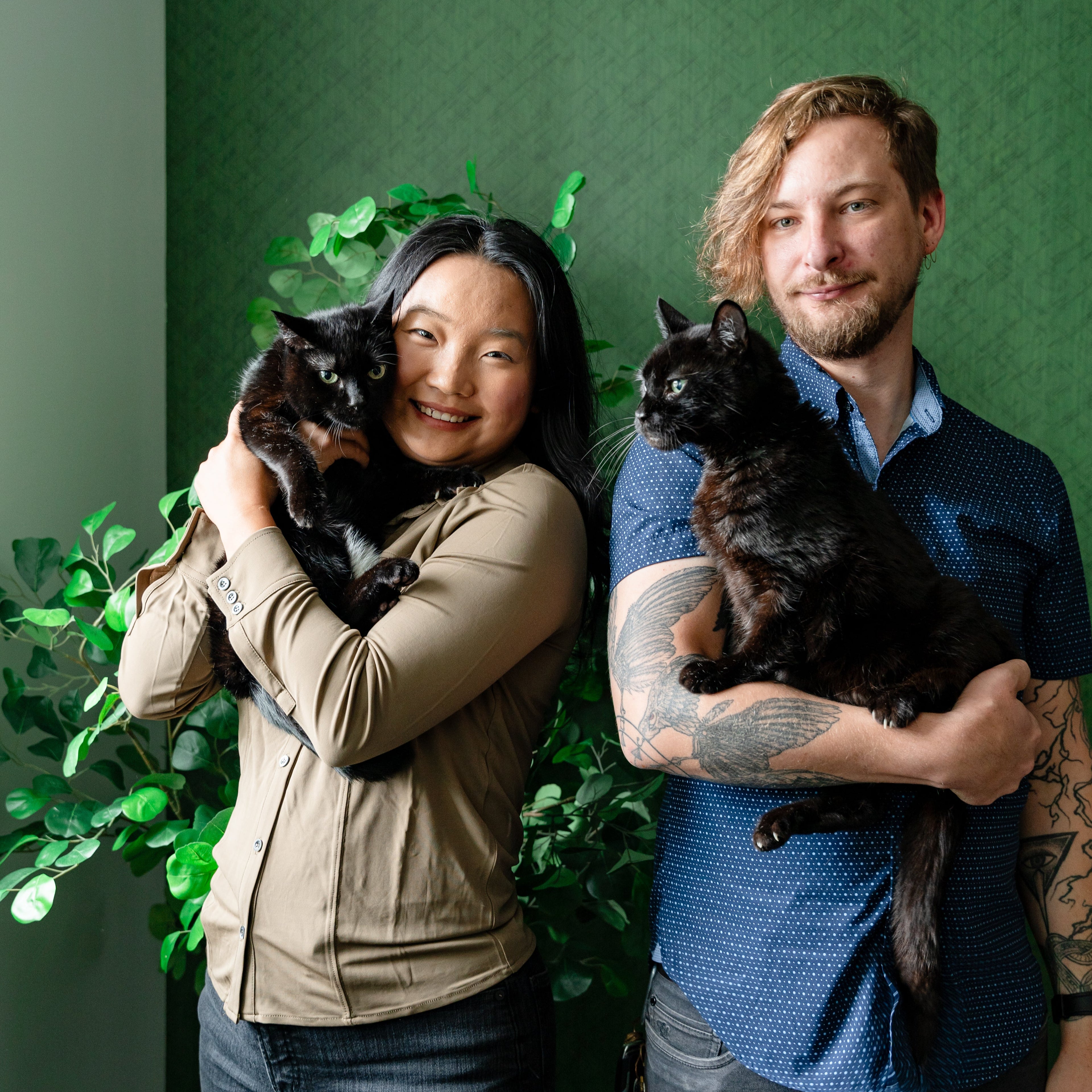 Two people holding black cats against a green wall with plants.